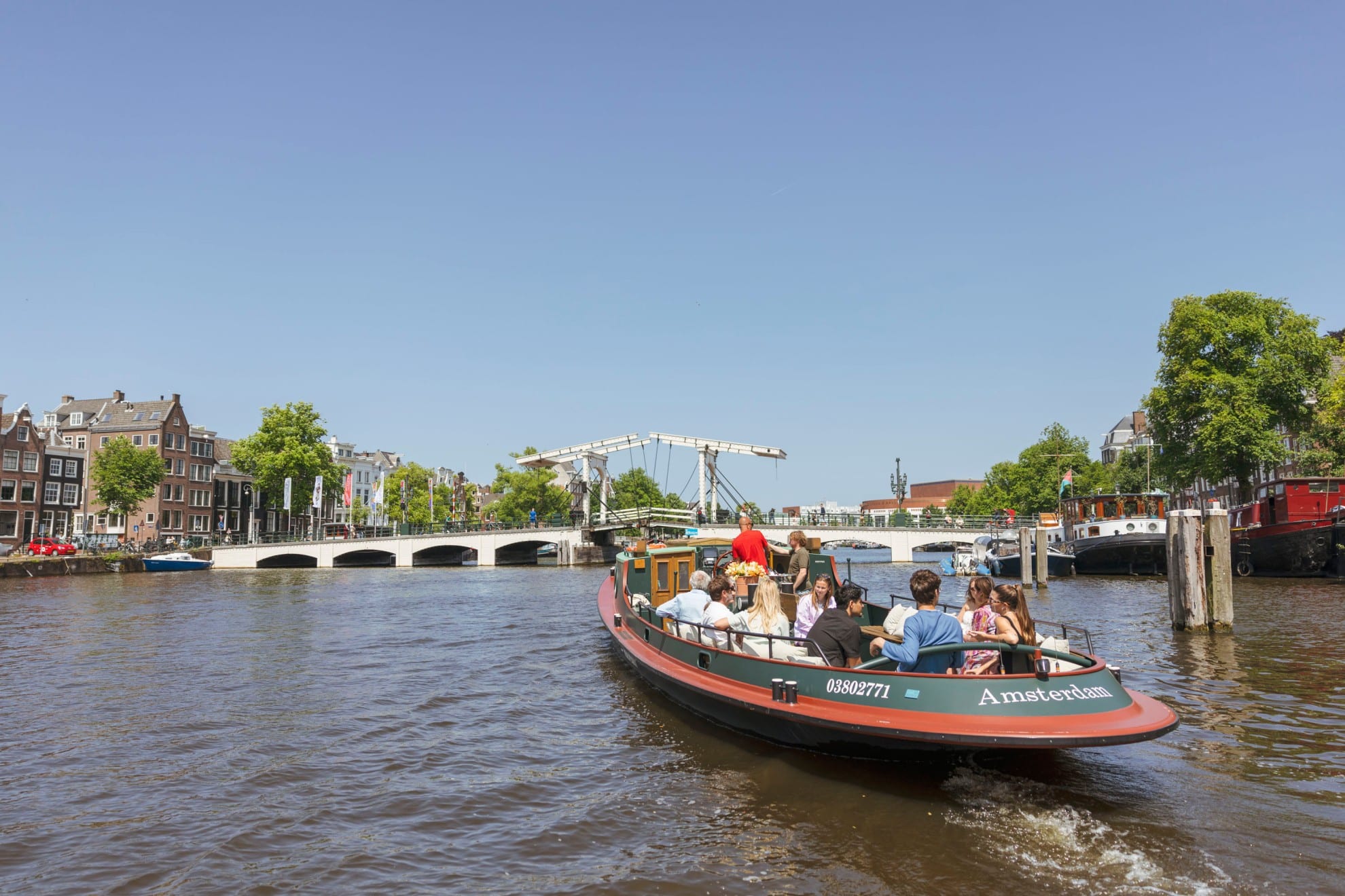 Ein klassisches Rundfahrtboot voller Passagiere fährt auf einer Amsterdamer Gracht Richtung Magere Brug an einem sonnigen Tag.